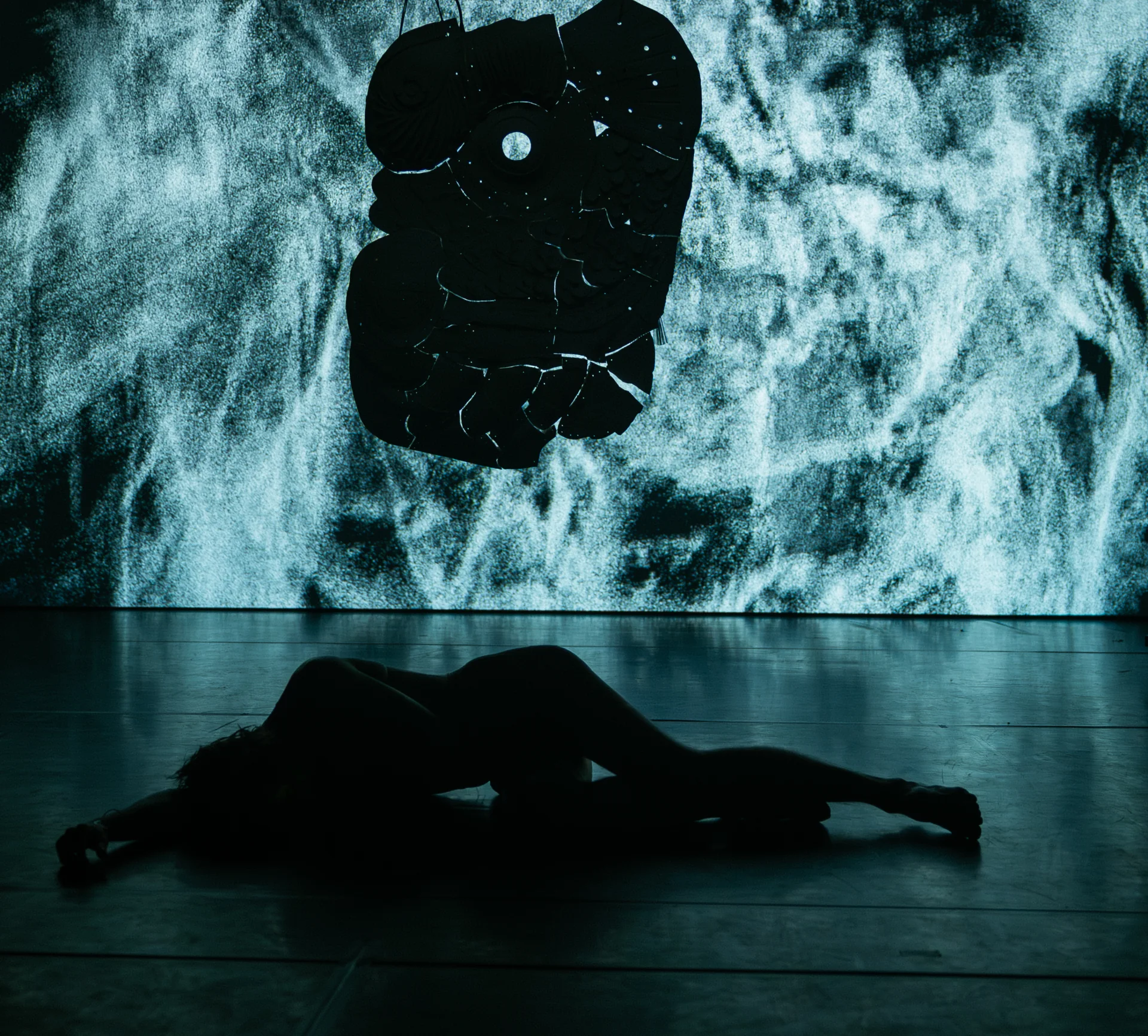 A body lies below a suspended ceramic object and a pale projection.
