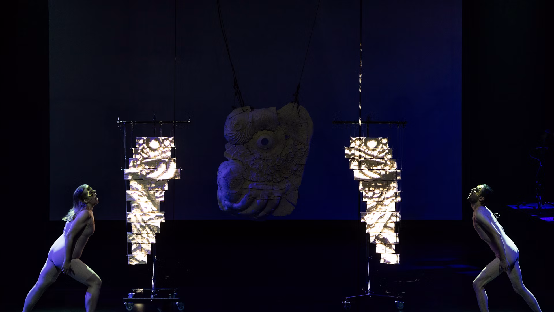 Two performers frame the stage between ceramic objects and projected images.